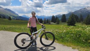Roxy standing roadside in full cycling kit with mountain bike in front of a sunny mountain range
