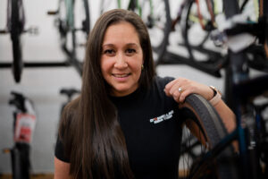 woman in a bike shop holding a rear wheel with a rack of bikes on the wall behind