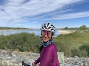 Woman in a cycling helmet, sunglasses and jacket smiling towards the camera with a blue sky and grassy beachfront in the background