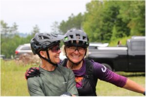 2 women smiling arm in arm in their mountain bike jerseys and helmets
