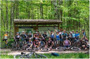 A group photo of women in the mtb exchange sitting in front of an information board in a forest