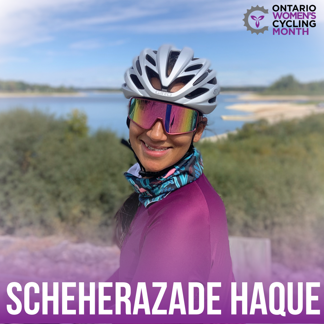 Woman in a cycling helmet, sunglasses and jacket smiling towards the camera with a blue sky and grassy beachfront in the background