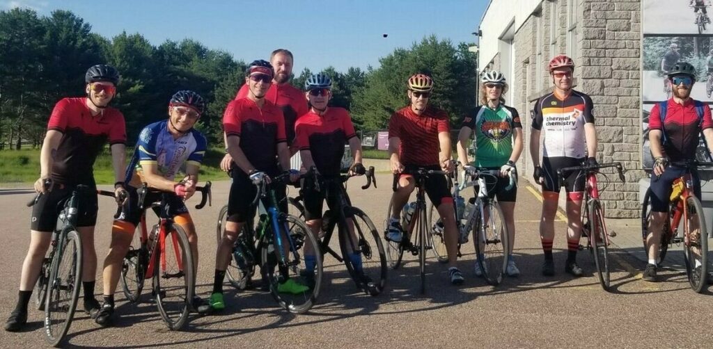 Group of GearHead Cycling Club cyclists standing beside a brick building smiling